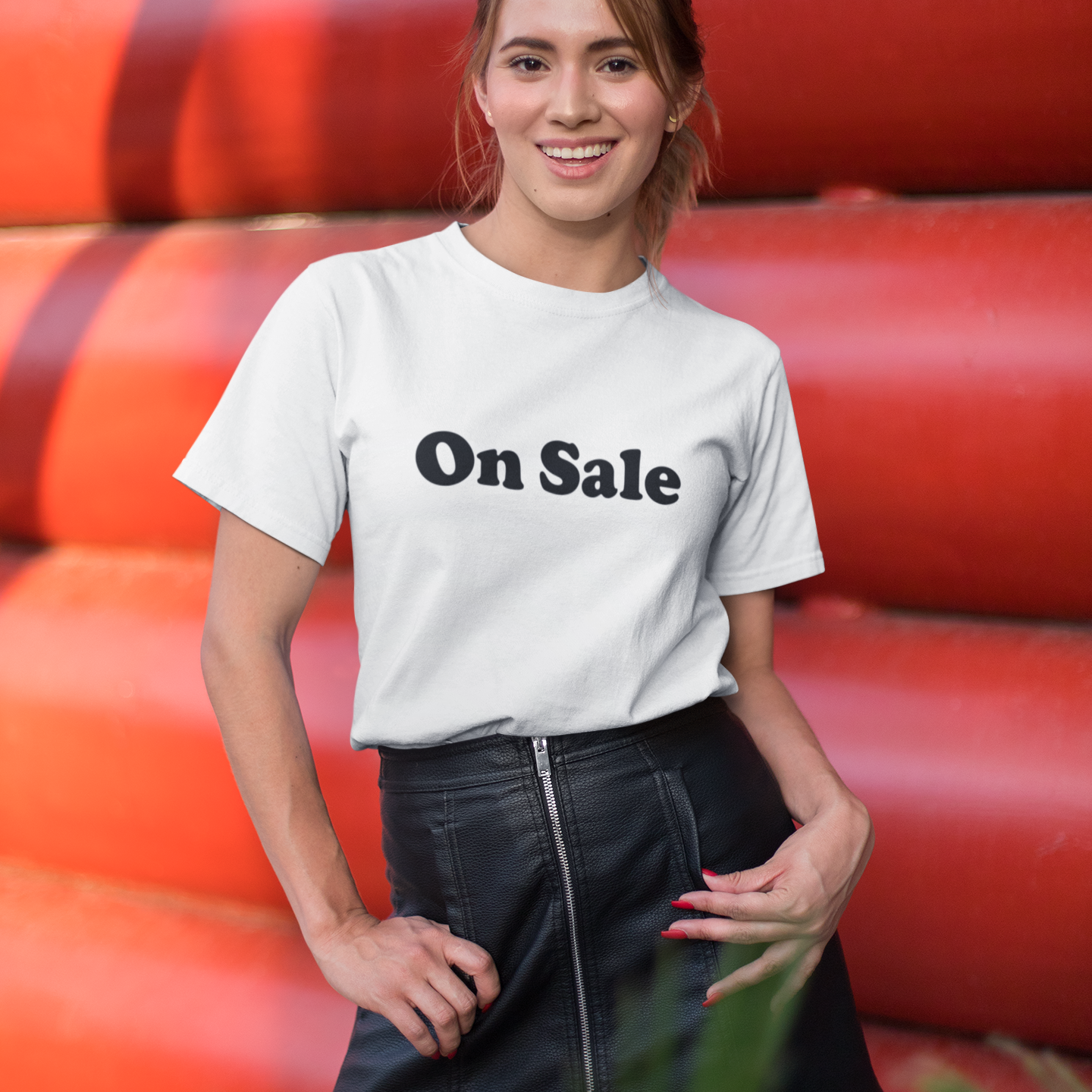 A woman is standing in front of a bold red background, smiling confidently. She is wearing a white t-shirt with the phrase "On Sale" printed in large black letters, paired with a high-waisted black leather mini skirt featuring a front zipper. Her relaxed pose and cheerful expression give off a stylish and playful vibe, while the eye-catching shirt adds a fun promotional element to the scene. Some greenery is visible in the foreground, adding a touch of natural contrast
